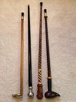 Four handcrafted walking sticks placed side by side on light carpet, showing varied handle designs and wood finishes.
