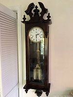 Full view of tall handcrafted Vienna regulator wall clock mounted on wall, showing wooden case, glass front, pendulum and weights.