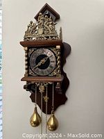 Front view showing ornate cast brass top with classical figures, silvered dial with Roman numerals, twisted brass columns and two pear-shaped weights hanging by chains.