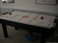 Air hockey table in a room showing the full length, playing surface, mallets, and pucks with power cord visible.
