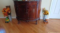 Front view of the carved half round antique cabinet with glass top and carved claw feet, placed against wall with hardwood floor.