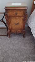 Photo of wooden antique nightstand with two drawers and carved decorative details, doily on top.