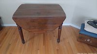 Wooden antique drop leaf table with one drop leaf raised and one lowered. Table has visible grain and carved turned legs. Photo taken to show table top and leg detailing.