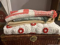 Four blankets stacked: coral and white crochet, green with fringes, beige fabric, white crochet with red flower motifs