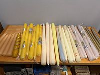 Full view of multiple candles in neutral colors arranged on wooden surface including sets wrapped in plastic and individual units of different shades and shapes.