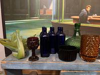 Photo of seven blue glass jars, green decanter, amber vessel, purple bowl, and red goblet showing variety of coloured glass art.
