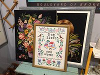Three fiber art cross stitch embroidery pieces displayed together, two framed and one unframed, showing floral designs and a 'God Bless Our Home' piece.