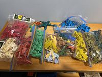 Photo showing multiple sealed and loose plastic toy figures in various colors including red, blue, yellow, green, brown, black, and white, arranged in ziplock and other clear bags on a surface