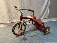 Side view of the entire red metal Radio Flyer tricycle showing the large front wheel, two smaller rear wheels, red plastic pedals, and white plastic seat.