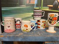 Seven mugs lined up on a blue shelf showing diverse colors and designs