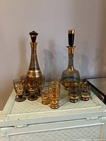 Two vintage 70s glass decanter sets displayed with accompanying shot glasses on woven white surface. One decanter is brown smoke with gold details and the other black smoke with gold details, all with original toppers.