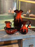 Four pieces of unmarked vintage Blue Mountain style pottery with bright orange and black glaze arranged on rustic table. Tall ruffled vase with scalloped rim stands in back, flanked by three smaller pitchers and a handled sugar bowl or creamer style vessel.