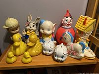 Front view of the full lot showing all vintage toys arranged on a wooden shelf including rubber ducks, clown toy, figurines, piggy banks and rooster squeeze toy.