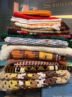 Stack of 16 vintage linen pieces with varied textures and colors including crocheted large tablecloth at bottom and woven carpets in the middle