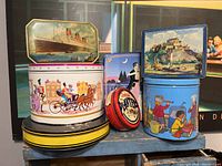 Overview photo showing all seven tins stacked and arranged in various shapes and sizes, with colorful vintage artwork including ship, dancing couple, children playing, castle, and Planters peanut tin.