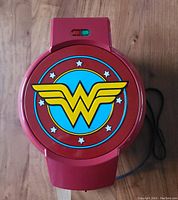 Top view of closed red waffle maker featuring colorful Wonder Woman logo on lid with indicator lights.