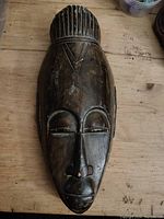 Front view of dark stained carved wooden tribal mask showing facial details and patterned hair or headdress.