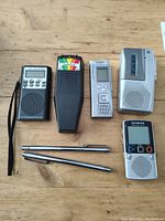 Photo of ghost hunting equipment including black ETC Research Device, EMF meter, Panasonic voice recorder, cassette player, Olympus voice recorder, and three metal antennas on a wooden surface.