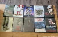 Lot of 10 horror DVDs laid out on wooden floor showing front covers including Jigsaw, IT, Beetlejuice, Saw series, Evil Dead 2, Land of the Dead, Winchester, Paranormal Activity 4, and Shutter Island.