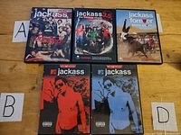 Top-down photo of the 5 Jackass DVDs aligned on wood floor with cards labeled A and B visible near two groups of cases