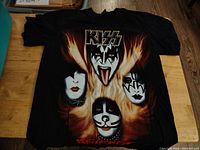 Front view of the black Kiss Revenge t-shirt showing the band's four members faces with flame graphics and logos.