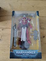Front view of the boxed McFarlane Warhammer 40,000 Adepta Sororitas Battle Sister action figure in red and white armor with accompanying weapons visible.