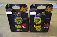Front view of two boxed Pokemon Pikachu LCD watches showing yellow plastic cases, printed bands, slide-on Pikachu charm, and packaging with Pokemon branding.