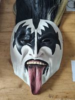 Front view of vintage Gene Simmons mask with Demon makeup design, showing detailed white rubber face, black paint accents, teeth, and extended tongue with black and white wig attached.