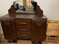 Front view of antique buffet cabinet showing dark stained wood, rectangular vintage mirror with scalloped wood backing, three central drawers with wooden handles, and two side doors with decorative carved wood embellishments. Cabinet stands on bun feet and shows polished finish.