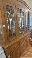 Front and side view of large wooden china cabinet with glass front doors showing glass shelves with lighting and wooden drawers and cabinets below.