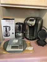 Photo of KitchenAid chopper with box, coffee brewer, digital kitchen scale, and mini waffle maker on kitchen counter.