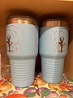 Two light blue Life Tree insulated cups with lids side by side on a floral tablecloth