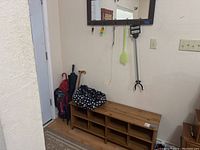 Overview of entryway showing wooden shoe caddy with multiple compartments at bottom, metal umbrella stand with umbrellas and walking cane beside it, wall mirror above with hooks