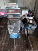 Image shows a grouped lot including a tripod, plastic storage boxes, a round clock, phone mounts, a solar eclipse kit box, and office supplies.