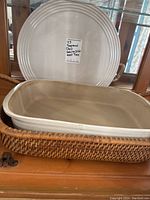 Photo showing large round ceramic tray placed vertically behind rectangular ceramic baking dish inside wicker basket on wooden surface.