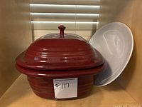 Photo showing rust-colored ceramic Dutch oven with lid and white oval platter behind it inside a cabinet.
