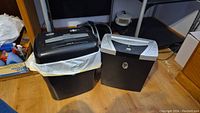 Photo showing two paper shredders side by side, one smaller Aurora with black body and silver top, one larger black Novitech shredder with silver top panel.