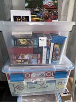 Two large plastic totes stacked. Top tote has visible puzzle boxes inside. Bottom tote is a Monopoly branded plastic case storing assorted games or puzzles.