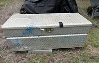 Front view of a large rectangular metal storage box with diamond plate texture, silver-gray color, blue paint marks, rust spots, and black strap securing lid