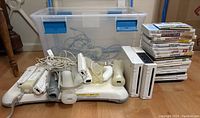 Photo of the entire lot showing two Wii consoles, multiple game cases, Wii accessories including remotes, nunchuk, balance board, and cables in a clear plastic container.