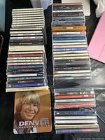 Photo of 66+ music CDs stacked in three piles over a black surface. Visible items include John Denver Greatest Hits, Cliff Richard 40 Golden Greats and various rock and classical compilations.