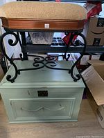 Photo showing stool with beige cushioned seat and wrought iron base sitting on top of a light green wood chest with metal latch and decorative molding, in storage area with other boxes around.