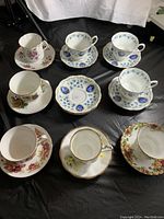 Overview of 8 cups and saucers plus 3 extra saucers displayed on black surface showing different floral designs and gold rims.