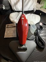 Red Dirt Devil Easy Steam Express II steamer with triangular black base, clear measuring cup, and instruction manual on a black surface, power cord visible.