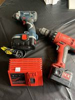 Two power drills (Milwaukee and Bosch) and a Milwaukee battery charger on a black surface.