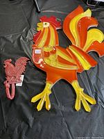 Large colorful lucite rooster with vibrant orange, yellow and red stripes and translucent material, laying flat on black surface next to smaller cast iron rooster wall hook.