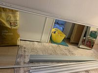 Photo shows one mirrored sliding door panel with white metal frame and attached rollers laying on a carpeted floor, with storage items in the background.