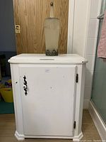 Full front shot of white wooden hamper cabinet with decorative handle and visible wear.