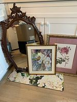 Full view of vintage carved wood mirror with floral carvings, leaning against white cabinetry alongside two framed floral artworks on the floor.