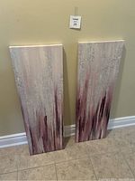 Photo of two acrylic paintings on canvas leaning against a beige wall on tiled floor. The paintings show vertical strokes and textures in white, pink, and dark burgundy tones.